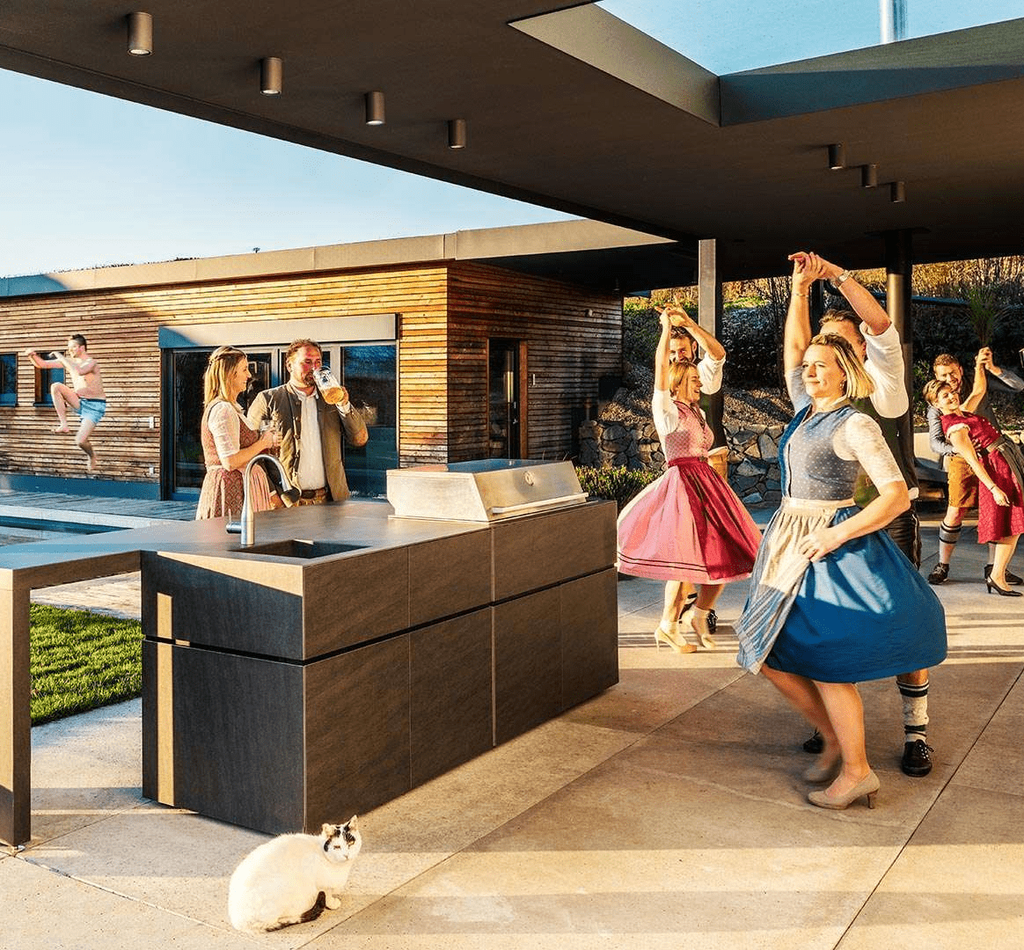 In Dirndl und Lederhosen gekleidete Gesellschaft, die auf einer Terrasse tanzt. Daneben eine Outdoorküche und im Hintergrund ein Pool