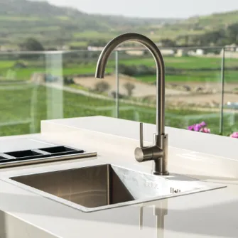 Spüle und Armatur einer Outdoorküche auf einem Balkon mit Glasgeländer und idyllisch grüner Landschaft dahinter