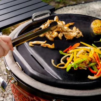 Nahaufnahme der Grillfläche eines Kamado Joe Grills auf dem mit einer Grillzange Fleisch- und Paprikastreifen gewendet werden