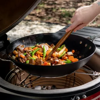 Gusseisenpfanne in einem Kamado Joe, in der mit einem Holzlöffel Gemüse und Fleisch gerührt wird