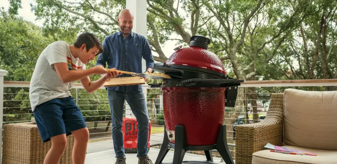 Ein Vater der ein großes Holzbrett bereit hält, damit sein Sohn die frisch gebackene Pizza aus dem Kamado Joe mit einer Pizzaschaufel darauf ablegen kann.